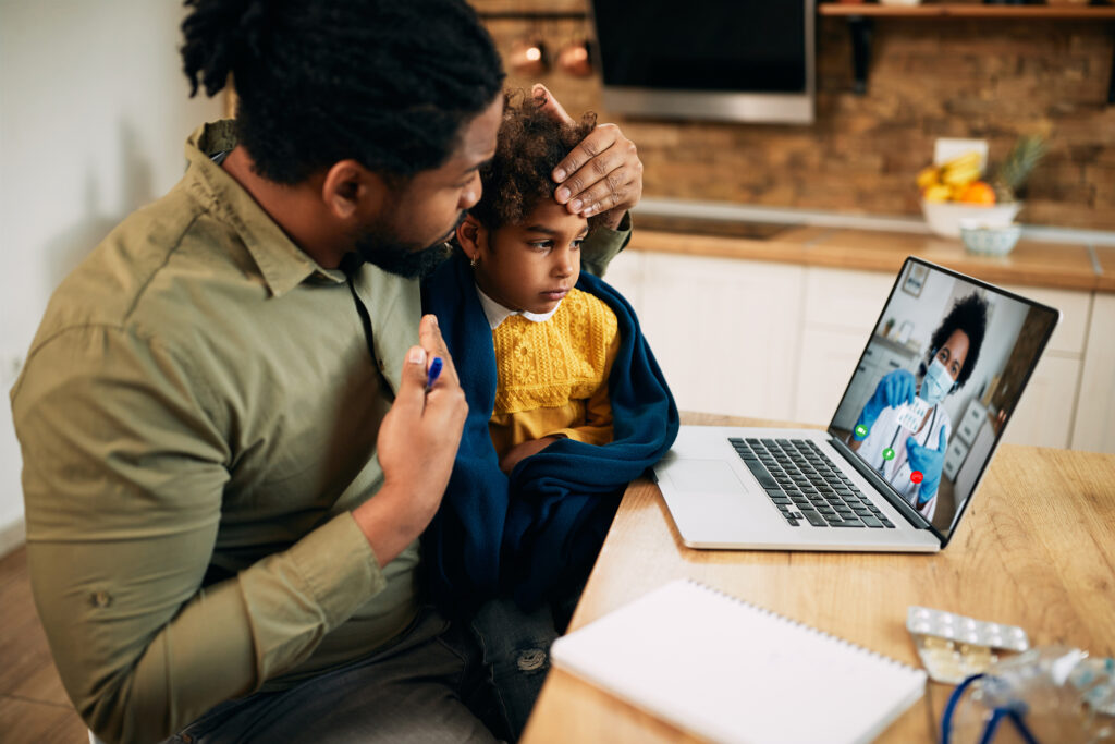 Father with child on lap in front of a laptop. Child appears ill.