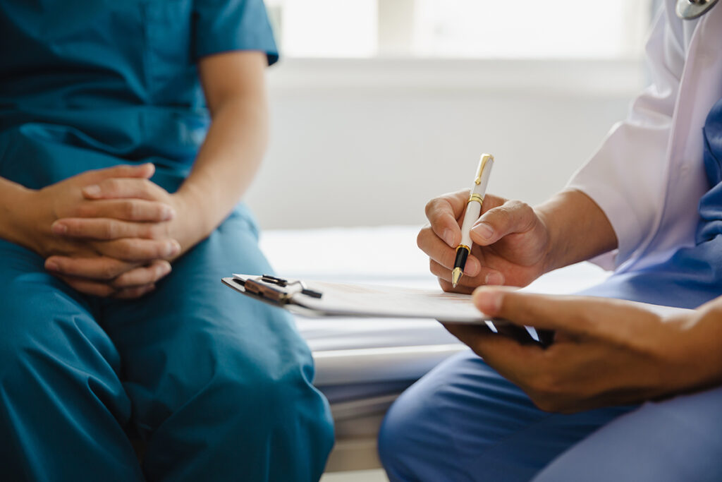 GP and person sitting on bed. GP with clipboard writing. Shot of body only.