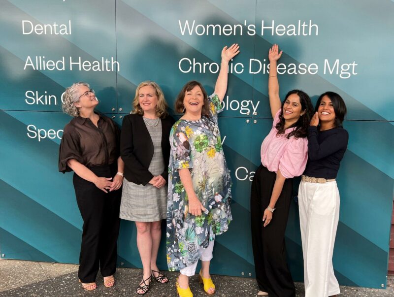 Five women standing in front of signage, pointing to women's health